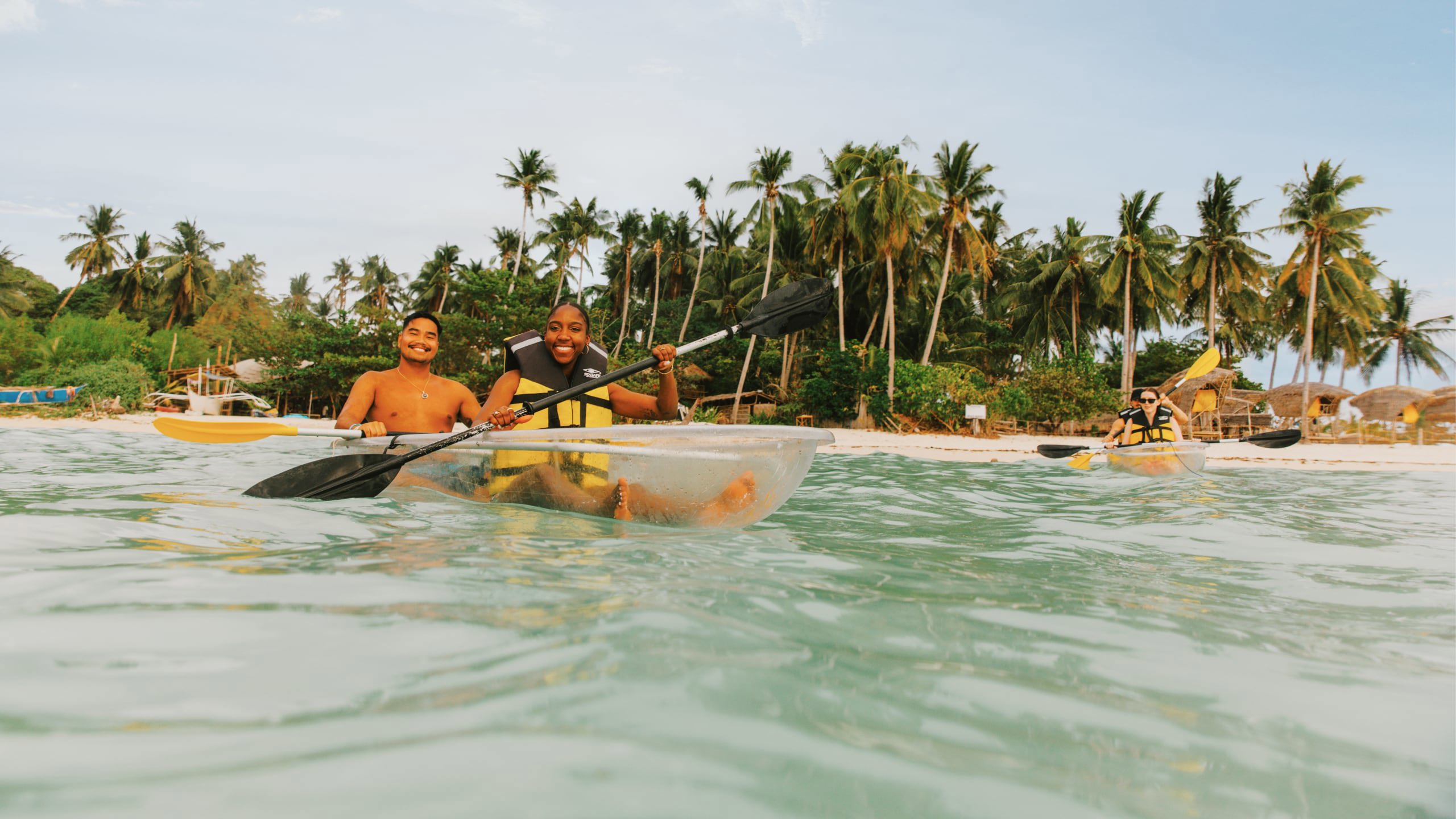 Two young travellers kayaking in a transparent kayak in crystal clear waters off a palm-lined beach in Palawan Philippines - Contiki Palawan Island Hopping Tour