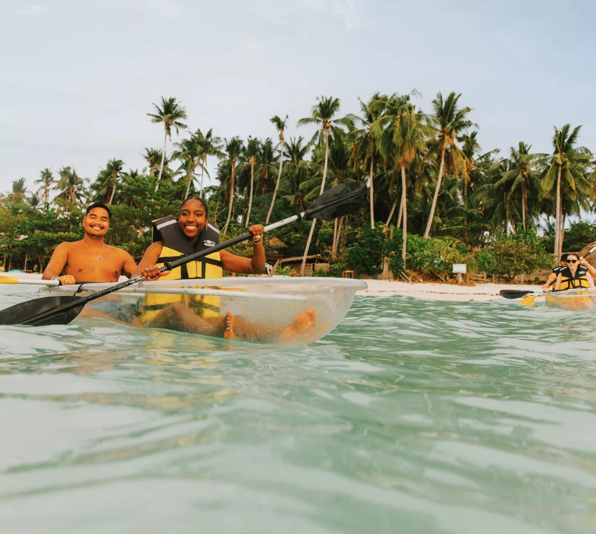 Two young travellers kayaking in a transparent kayak in crystal clear waters off a palm-lined beach in Palawan Philippines - Contiki Palawan Island Hopping Tour
