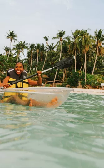 Two young travellers kayaking in a transparent kayak in crystal clear waters off a palm-lined beach in Palawan Philippines - Contiki Palawan Island Hopping Tour