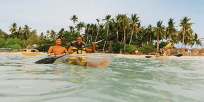 Two young travellers kayaking in a transparent kayak in crystal clear waters off a palm-lined beach in Palawan Philippines - Contiki Palawan Island Hopping Tour
