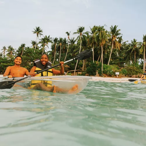 Two young travellers kayaking in a transparent kayak in crystal clear waters off a palm-lined beach in Palawan Philippines - Contiki Palawan Island Hopping Tour