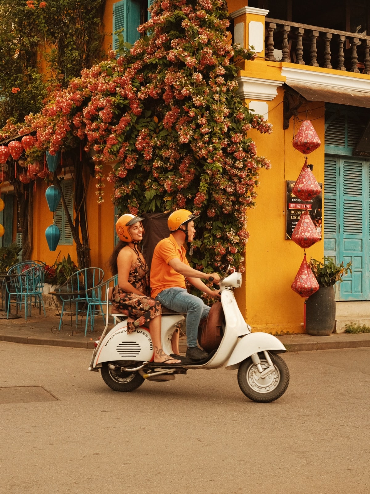 Travellers ride a vintage Vespa scooter past a flower-covered building and hanging lanterns in Hoi An, Vietnam.