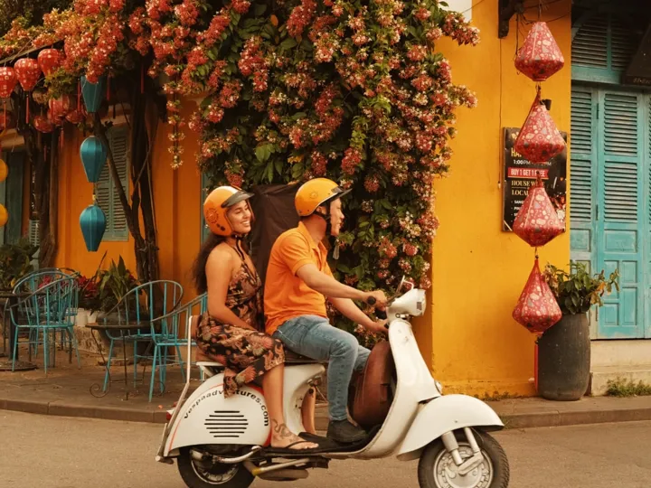 Travellers ride a vintage Vespa scooter past a flower-covered building and hanging lanterns in Hoi An, Vietnam.