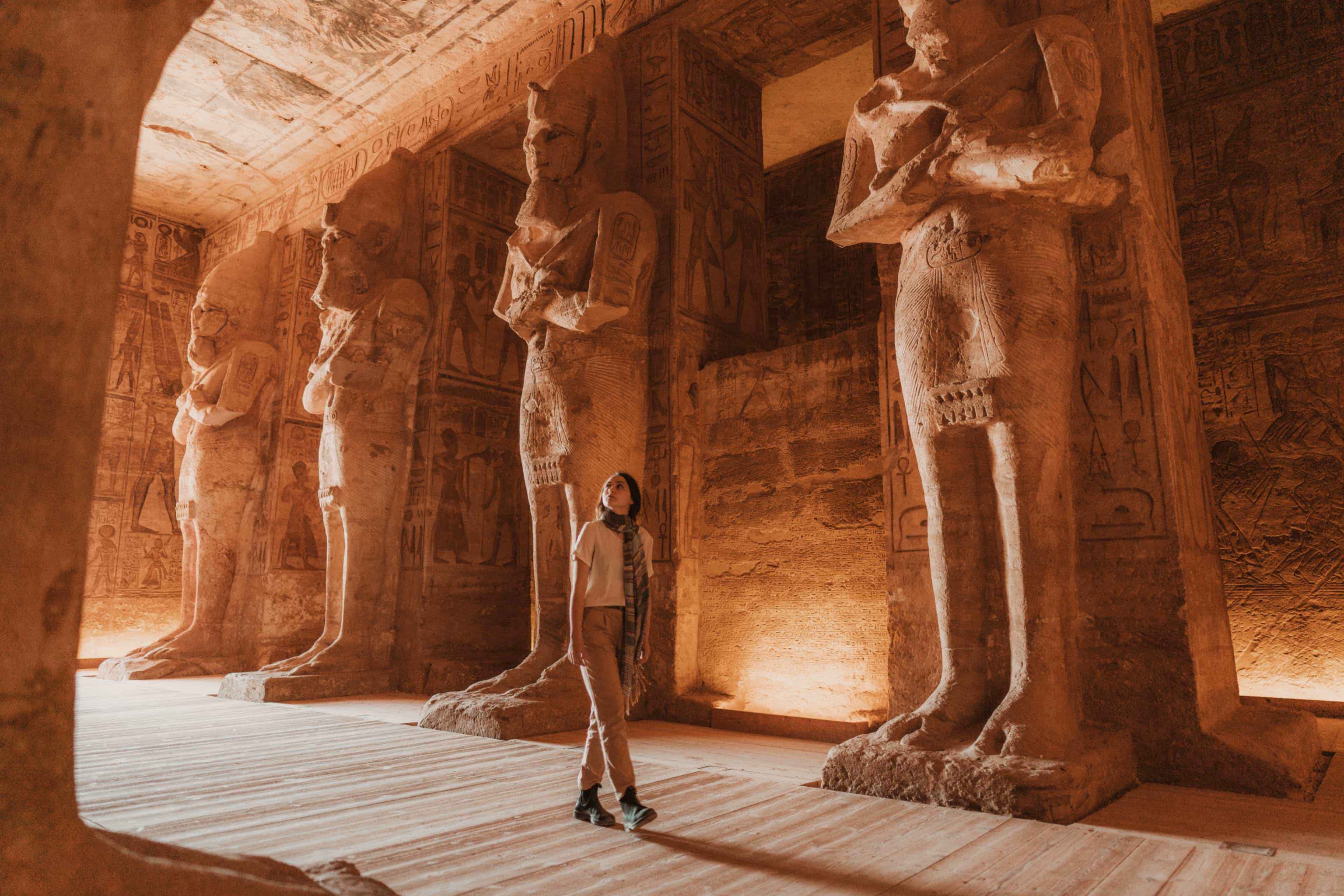 A traveller gazes up at towering carved pillar statues inside the hypostyle hall of Abu Simbel temple, illuminated in warm golden light.