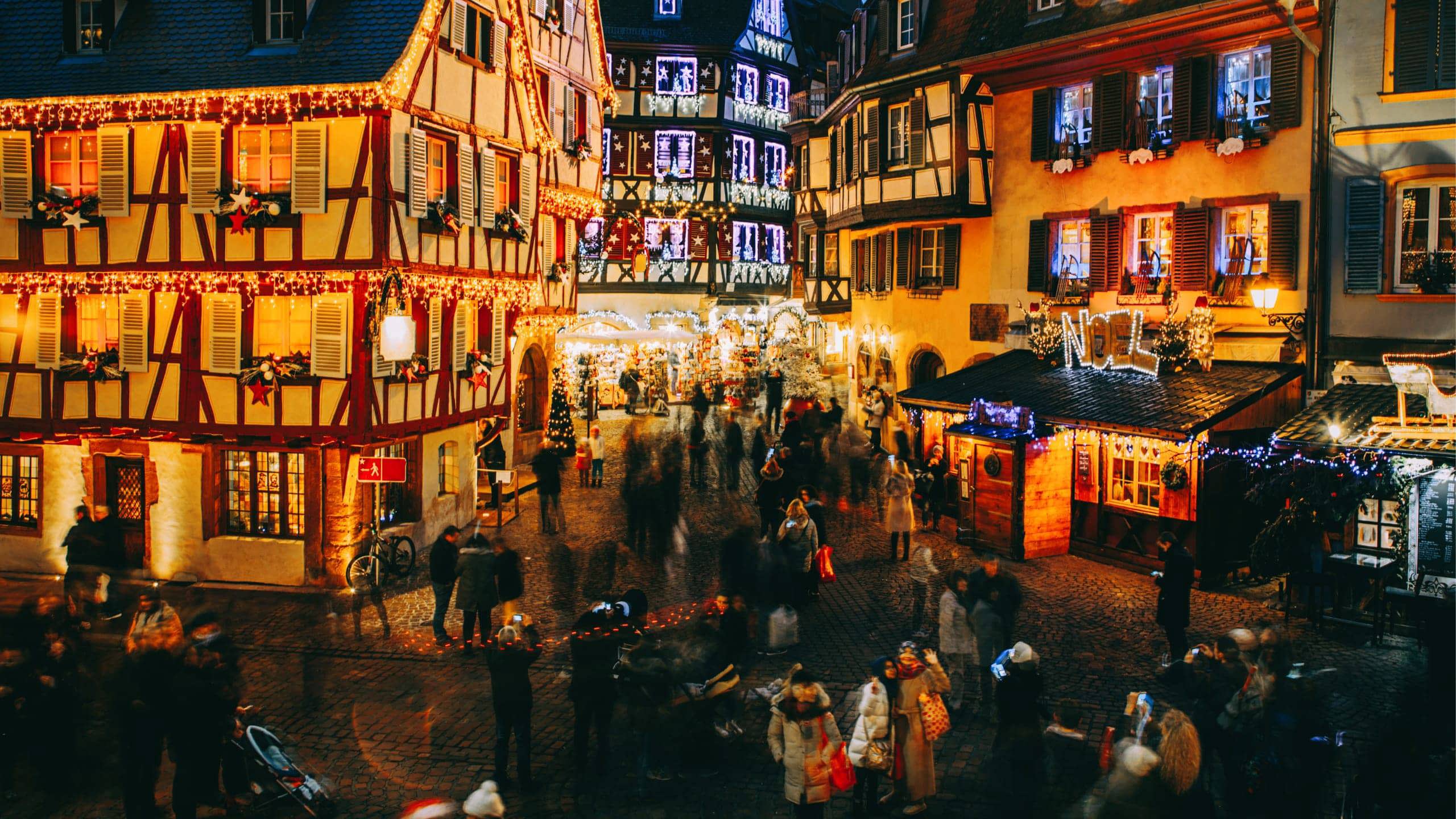 Aerial view of the festive Christmas market in France at night with illuminated half-timbered buildings and crowds of shoppers - Contiki French Christmas Markets Tour