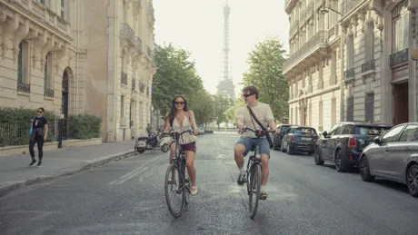Two travellers cycle down a tree-lined street in Paris with the Eiffel Tower rising in the background.