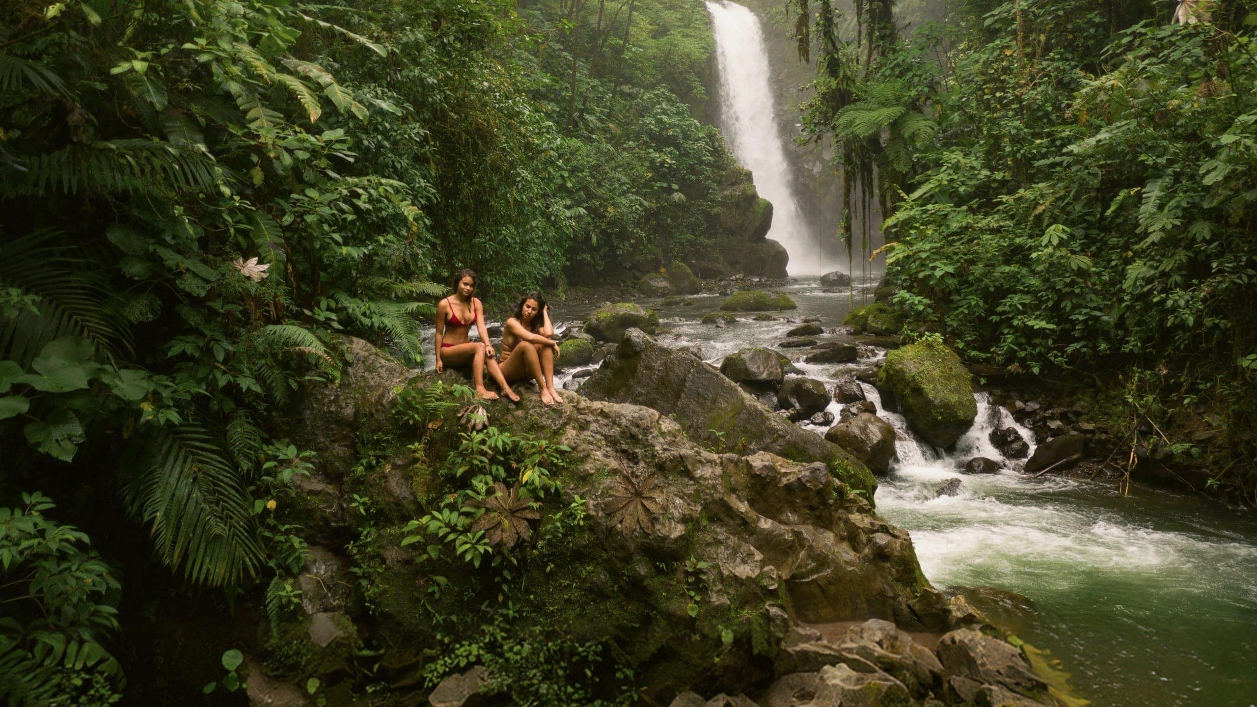 Two travellers relax on rocks beside a rainforest waterfall in Costa Rica, surrounded by lush jungle and a flowing river - Contiki Best of Costa Rica Tour