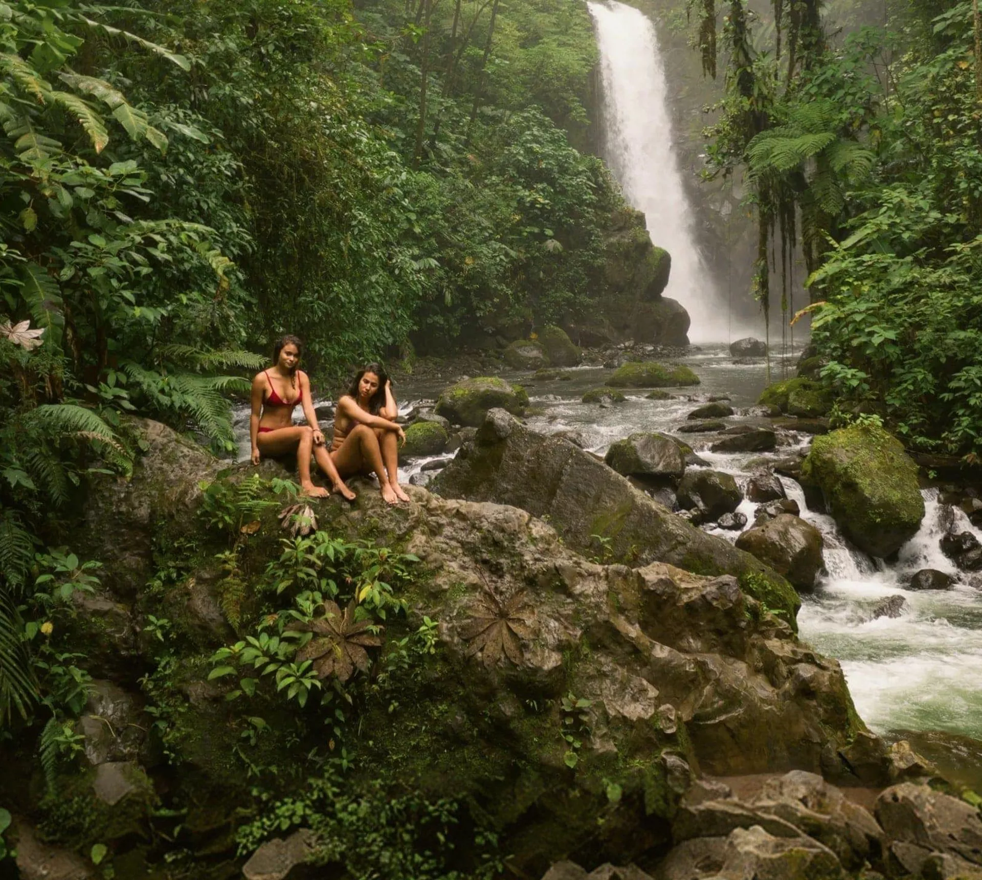Two travellers relax on rocks beside a rainforest waterfall in Costa Rica, surrounded by lush jungle and a flowing river - Contiki Best of Costa Rica Tour
