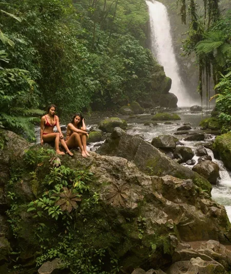Two travellers relax on rocks beside a rainforest waterfall in Costa Rica, surrounded by lush jungle and a flowing river - Contiki Best of Costa Rica Tour