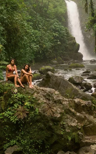 Two travellers relax on rocks beside a rainforest waterfall in Costa Rica, surrounded by lush jungle and a flowing river - Contiki Best of Costa Rica Tour