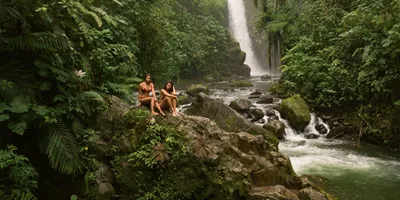 Two travellers relax on rocks beside a rainforest waterfall in Costa Rica, surrounded by lush jungle and a flowing river - Contiki Best of Costa Rica Tour