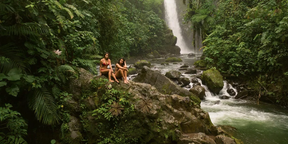 Two travellers relax on rocks beside a rainforest waterfall in Costa Rica, surrounded by lush jungle and a flowing river - Contiki Best of Costa Rica Tour