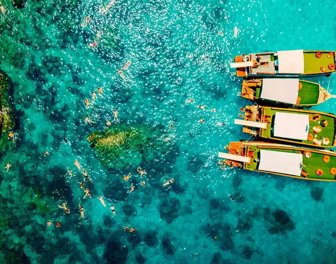 Contiki travellers swimming and snorkelling in vivid turquoise waters around traditional wooden boats anchored off the coast of a Thai island on a Thai Island Hopping East Tour