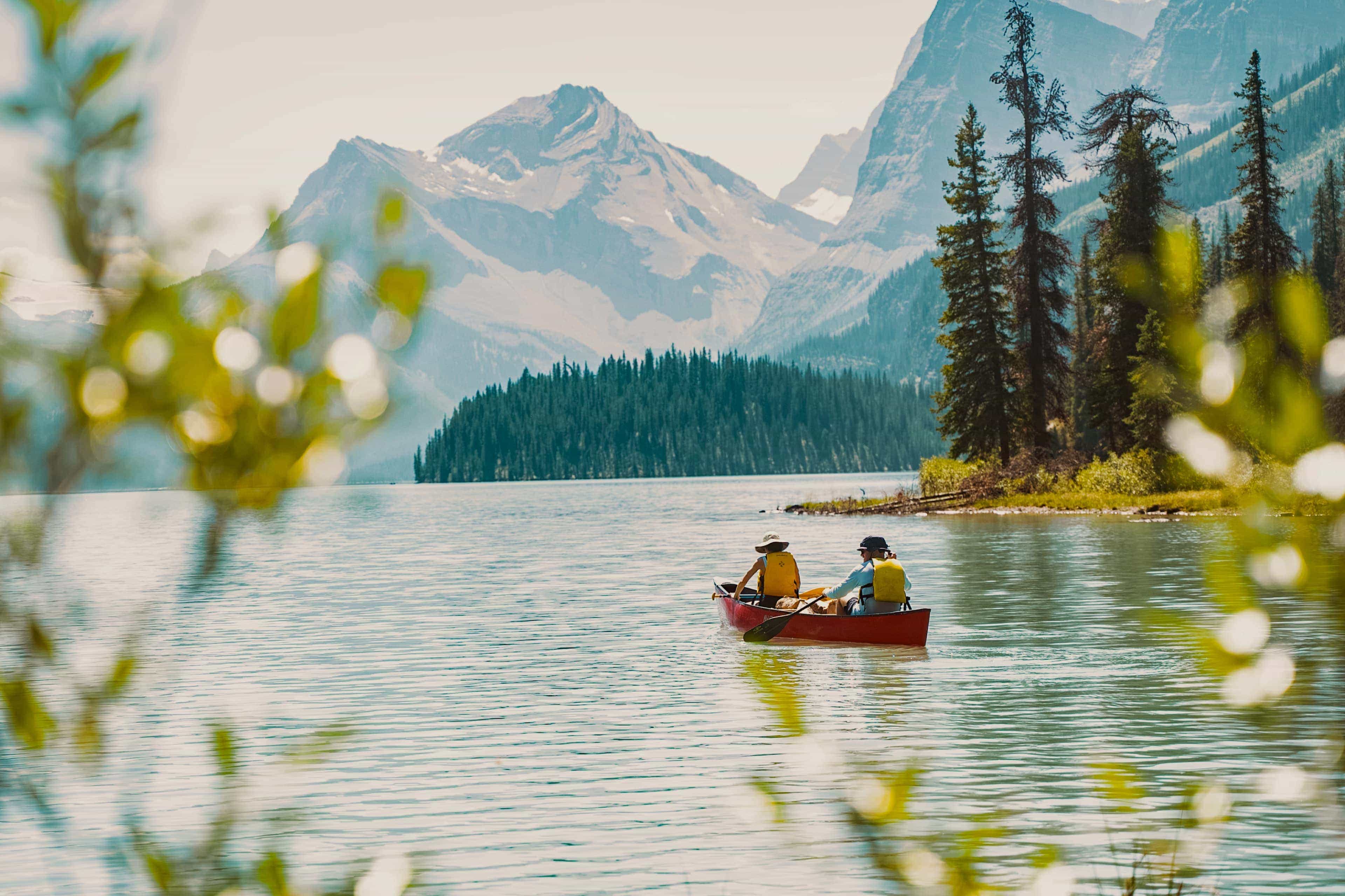 Two travellers canoeing across a calm mountain lake surrounded by pine forest and snow-capped peaks.