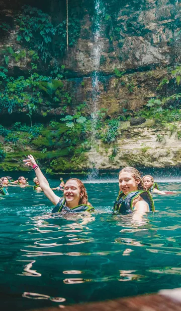 Two Women Swimming In A Natural Pool With A Group Of People