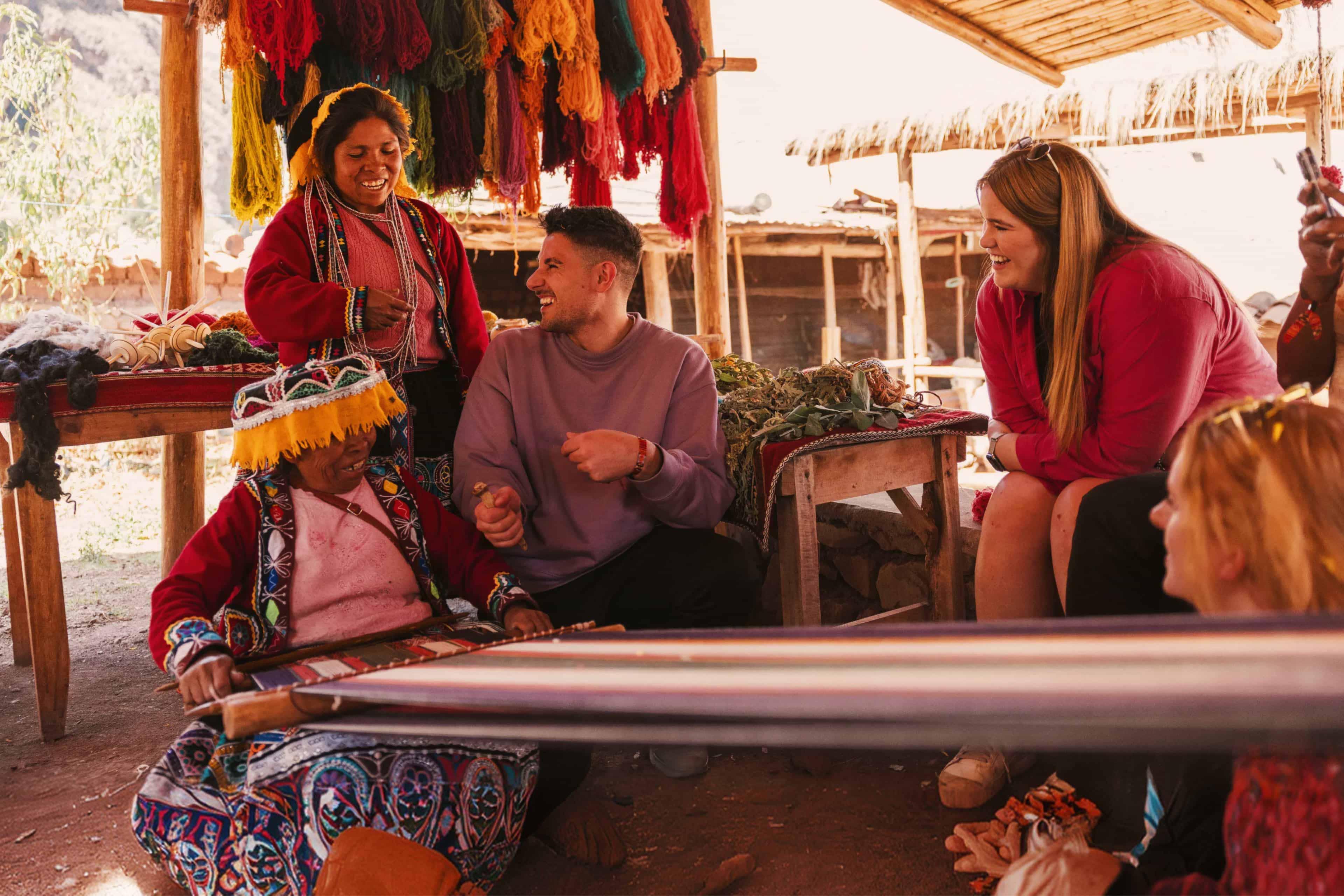 Travellers laughing with local weavers beneath dyed wool skeins at a textile workshop in Peru.