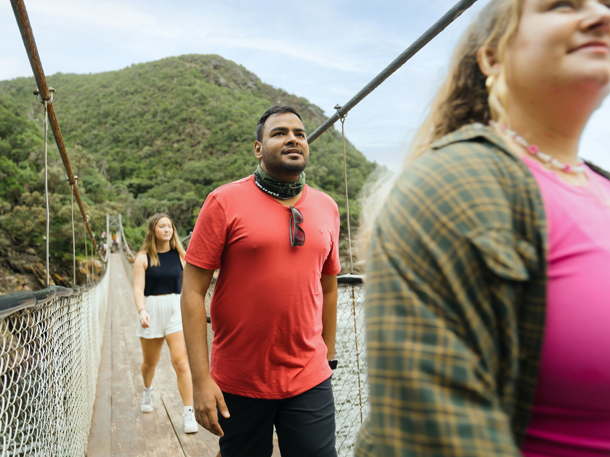 Three People Crossing A Narow Bridge Green Mountain At The Back