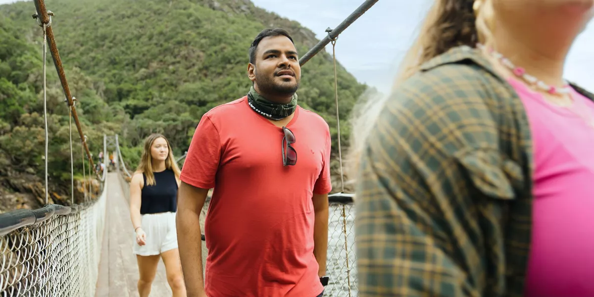 Three People Crossing A Narow Bridge Green Mountain At The Back