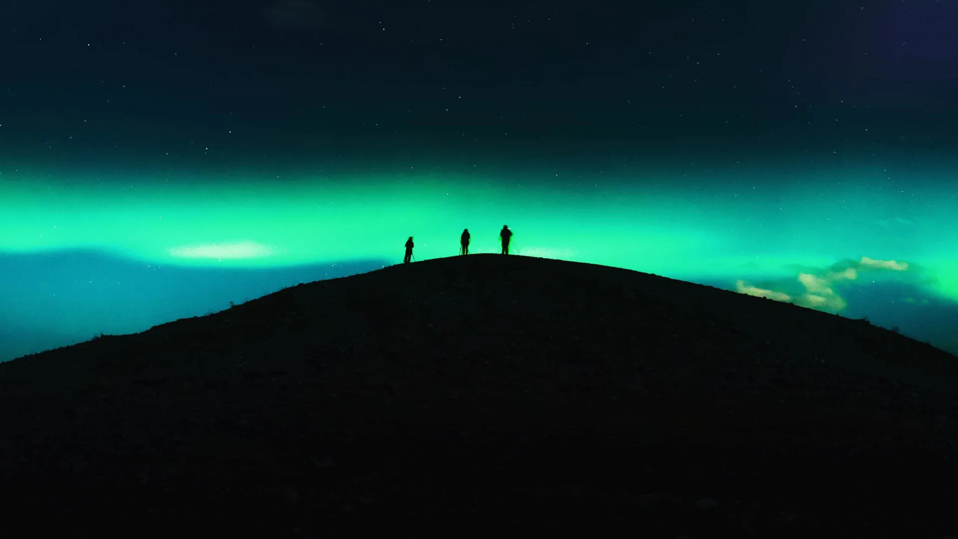 Three travellers silhouetted on a dark hilltop beneath a vivid green Aurora Borealis and starry night sky in Tromsø Norway - Contiki Norway Fjords and Northern Lights Tour