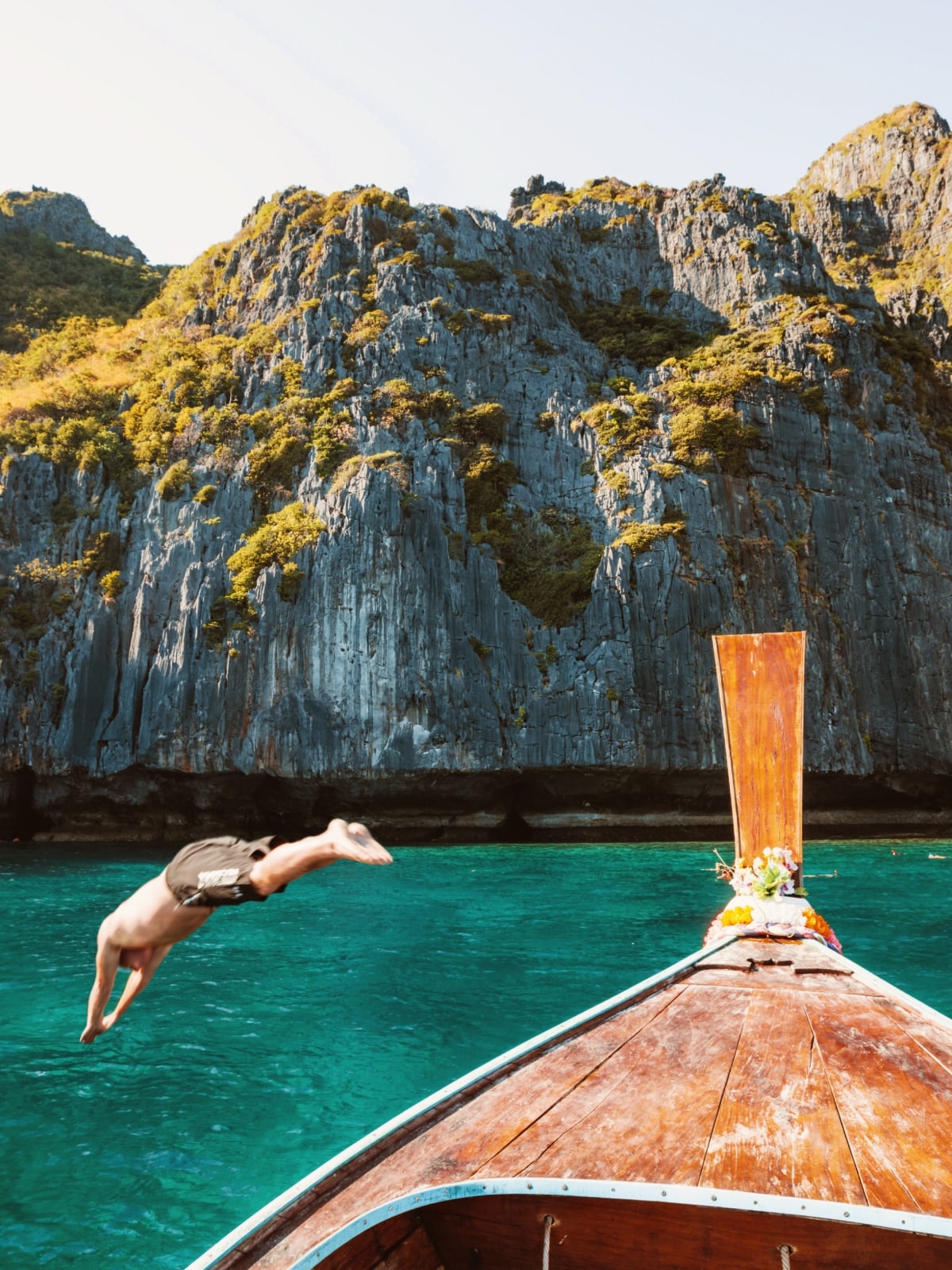 A traveller dives from a traditional longtail boat into turquoise water beneath towering limestone cliffs in Thailand.