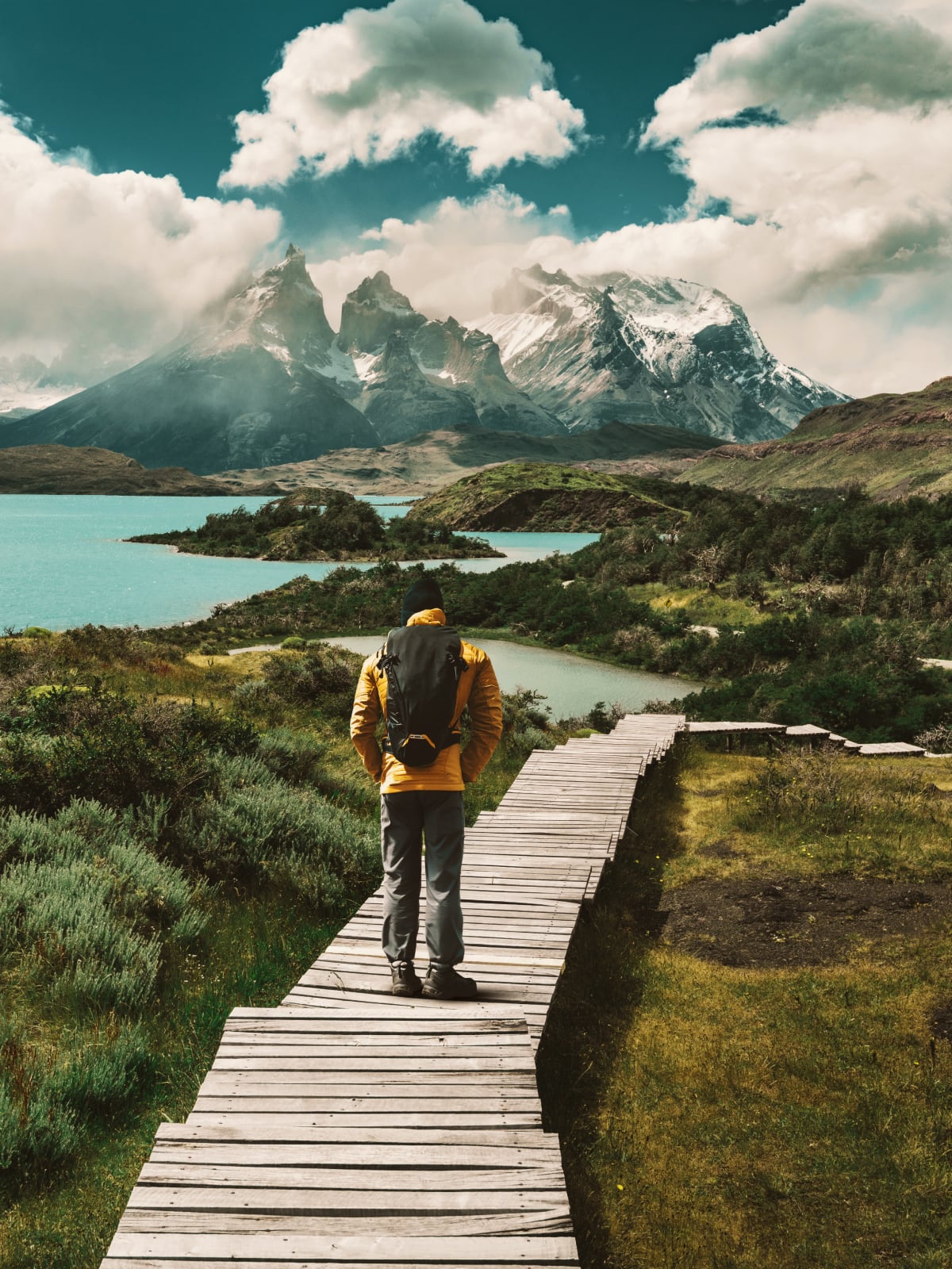 Hiker walking a wooden boardwalk toward the jagged snow-capped peaks of Torres del Paine, Patagonia.