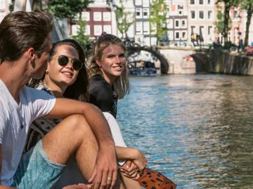 Contiki travellers relax beside an Amsterdam canal, with historic bridges and canal houses in the background on the European Escapade Tour