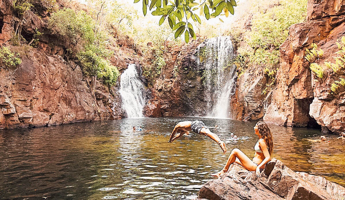 A woman sitting on the rocks and a man jumping into the water surrounded by plants and waterfalls