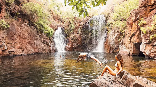 A woman sitting on the rocks and a man jumping into the water surrounded by plants and waterfalls