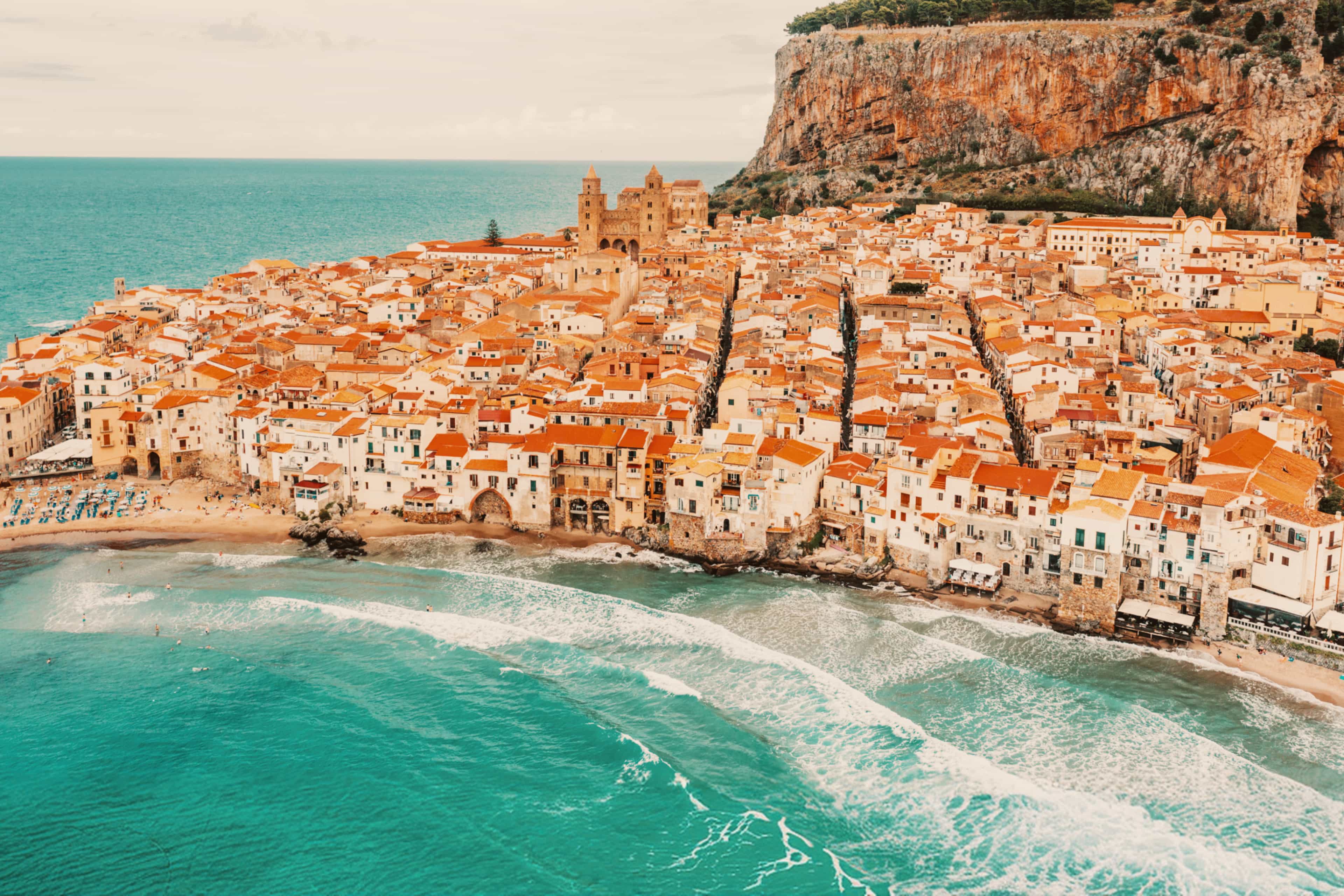 Aerial view of the coastal town of Cefalù, Sicily, with terracotta rooftops lining the beach beneath a rocky cliff.