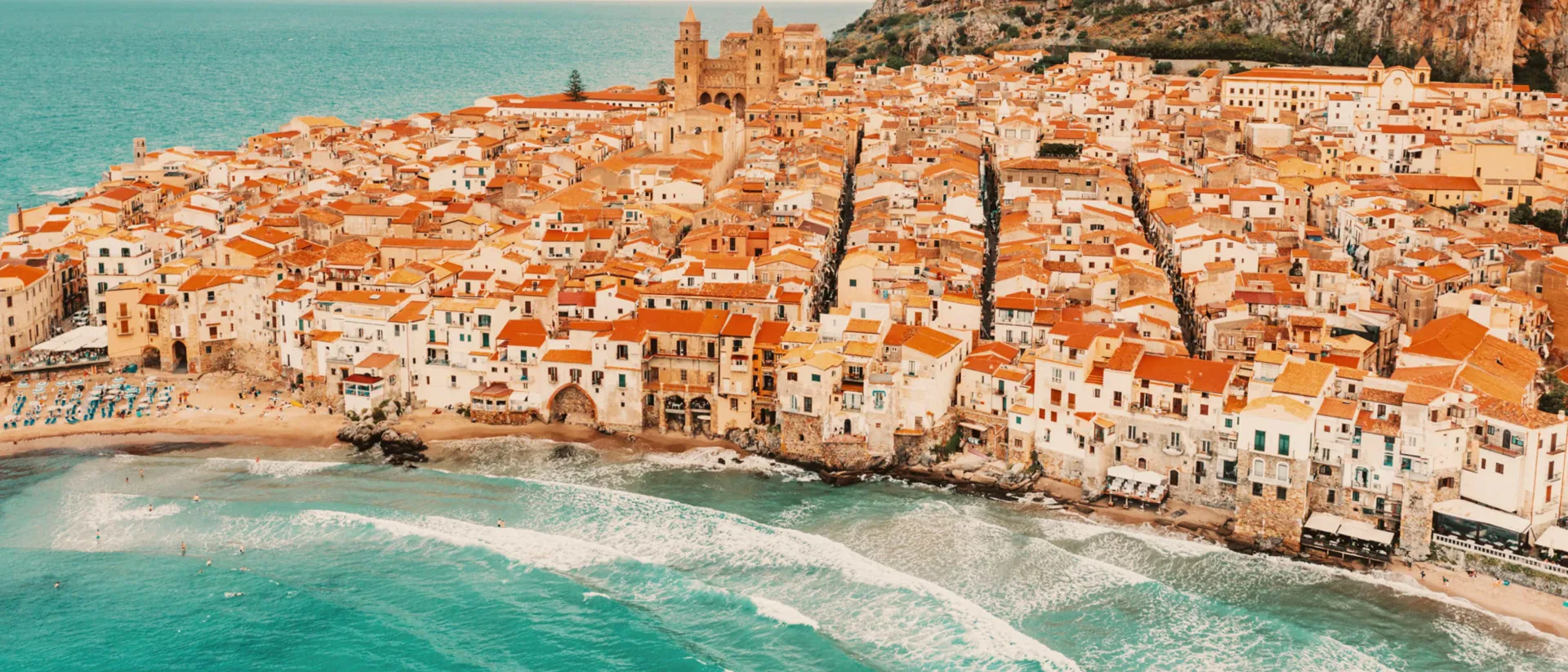 Aerial view of the coastal town of Cefalù, Sicily, with terracotta rooftops lining the beach beneath a rocky cliff.