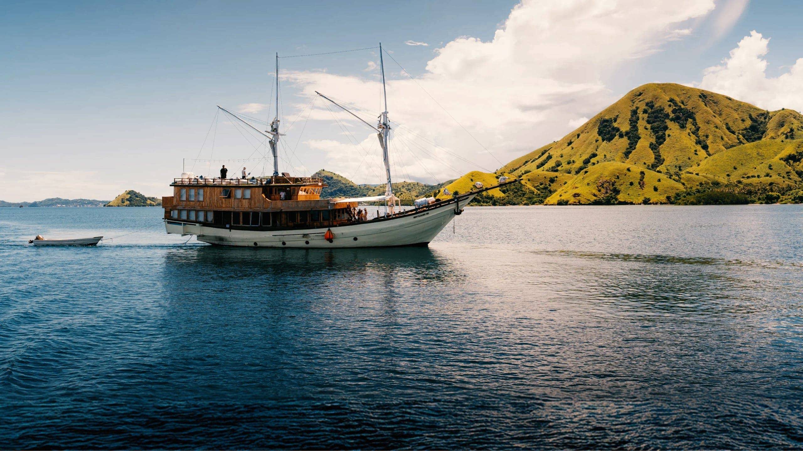 Traditional wooden phinisi sailing boat cruising through calm waters past the lush green hills of Komodo National Park Indonesia - Contiki Komodo and Flores Sailing Tour