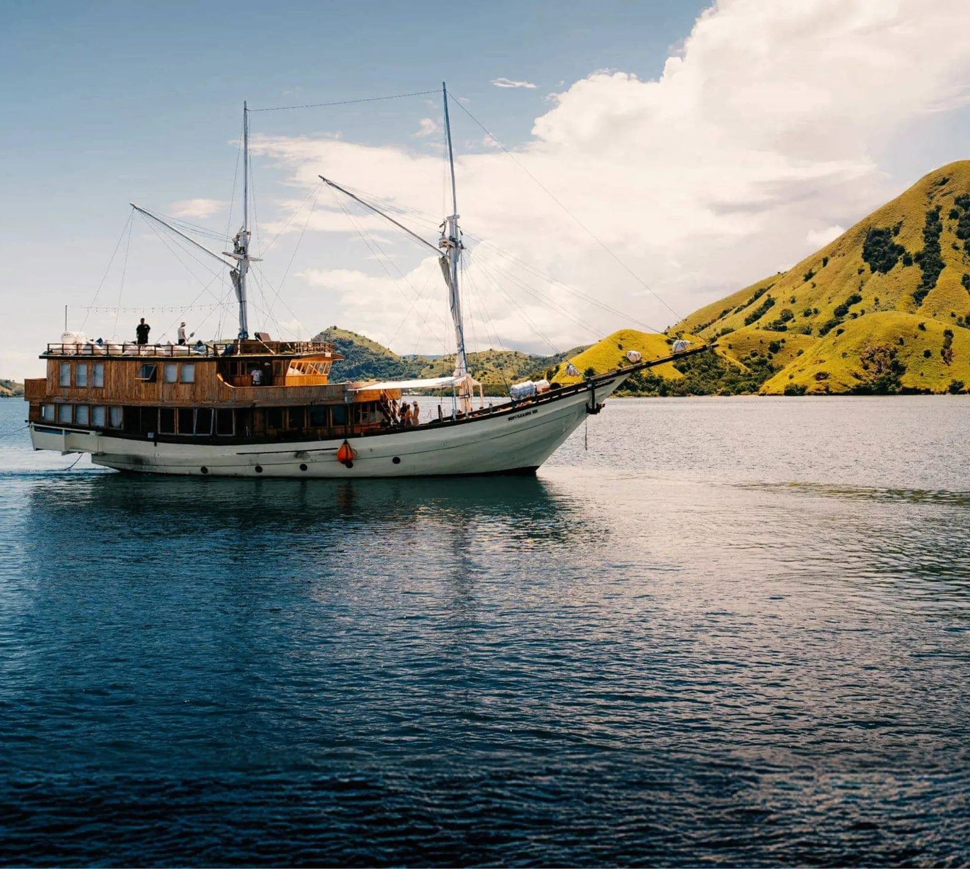 Traditional wooden phinisi sailing boat cruising through calm waters past the lush green hills of Komodo National Park Indonesia - Contiki Komodo and Flores Sailing Tour