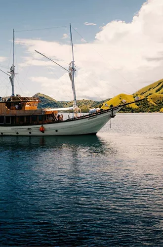 Traditional wooden phinisi sailing boat cruising through calm waters past the lush green hills of Komodo National Park Indonesia - Contiki Komodo and Flores Sailing Tour