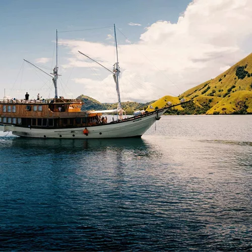 Traditional wooden phinisi sailing boat cruising through calm waters past the lush green hills of Komodo National Park Indonesia - Contiki Komodo and Flores Sailing Tour