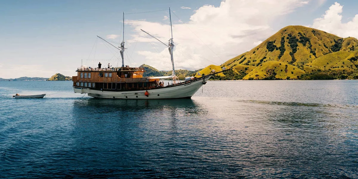 Traditional wooden phinisi sailing boat cruising through calm waters past the lush green hills of Komodo National Park Indonesia - Contiki Komodo and Flores Sailing Tour