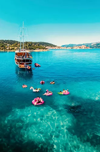 Drone view of travellers on colourful pool inflatables in clear turquoise water alongside a wooden gulet off the Croatian Dalmatian coast - Contiki Croatia Sail