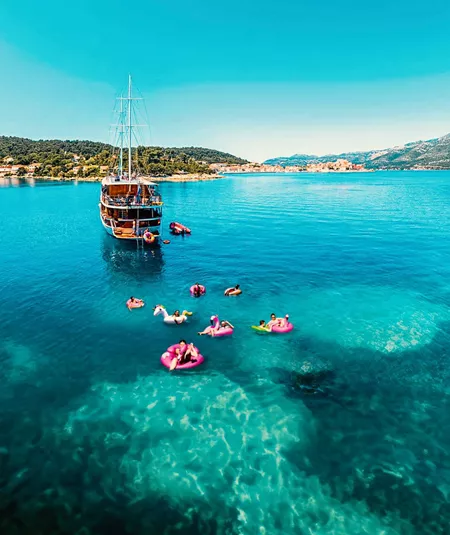 Drone view of travellers on colourful pool inflatables in clear turquoise water alongside a wooden gulet off the Croatian Dalmatian coast - Contiki Croatia Sail