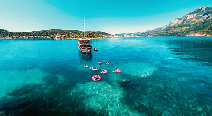 Drone view of travellers on colourful pool inflatables in clear turquoise water alongside a wooden gulet off the Croatian Dalmatian coast - Contiki Croatia Sail