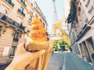 A freshly baked croissant held up towards the Eiffel Tower on a quiet Paris street during the Contiki European Horizon Tour