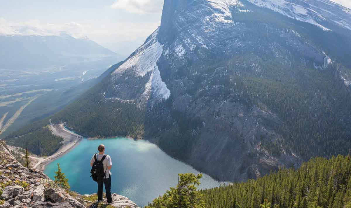 Man Standing At The Top Of A Mountain Looking At Other Mountains And Lake