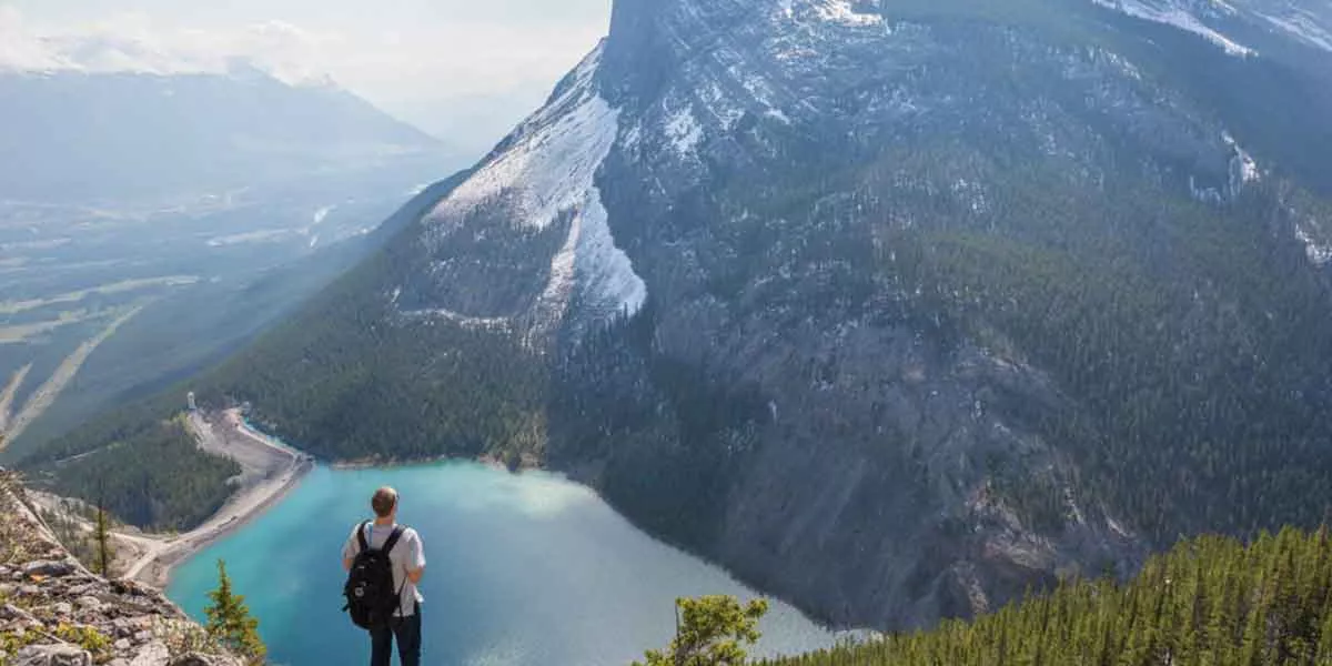 Man Standing At The Top Of A Mountain Looking At Other Mountains And Lake