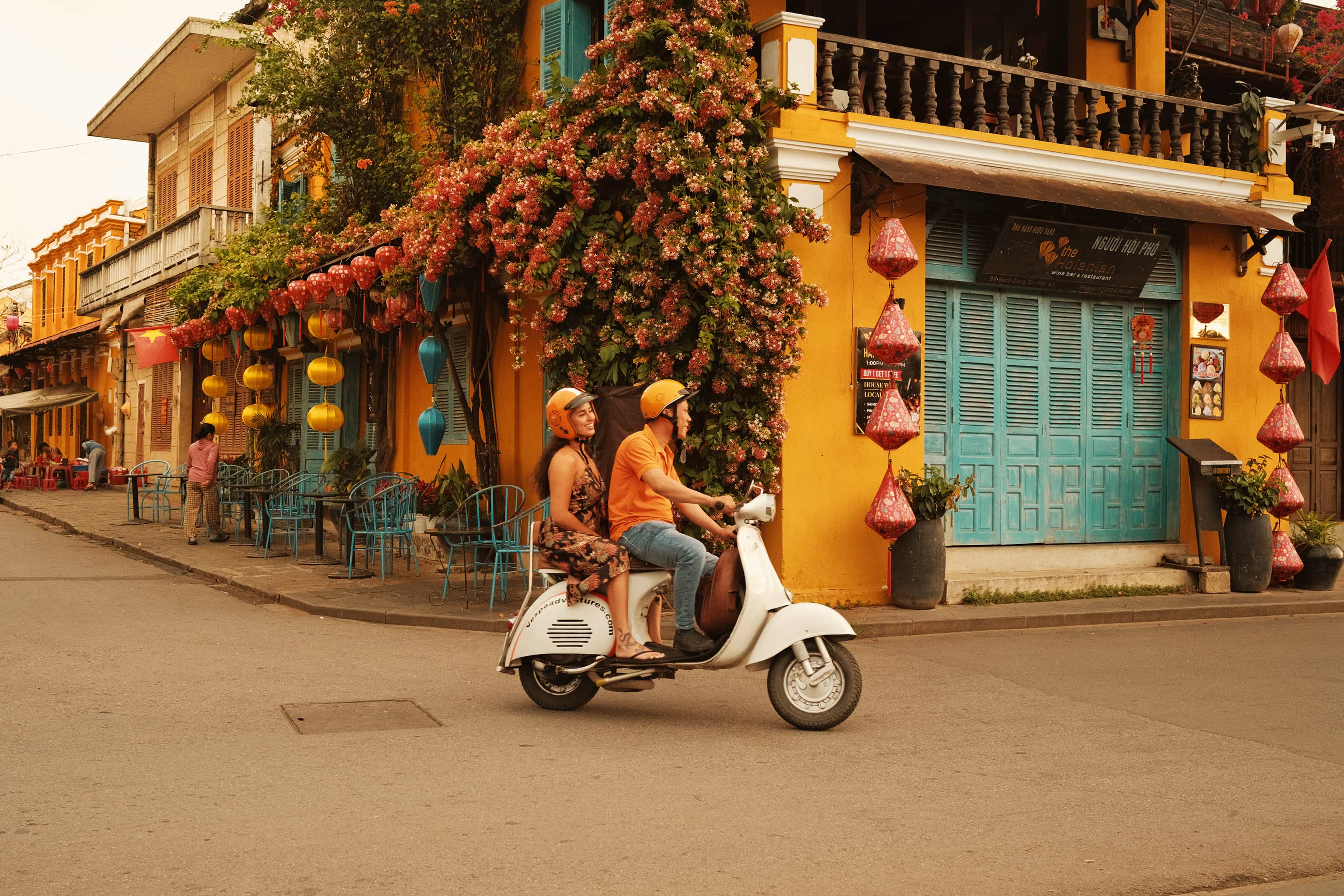 Travellers riding a scooter past flower-covered yellow buildings and lanterns in Hoi An, Vietnam.