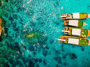 Contiki travellers swimming and snorkelling in vivid turquoise waters around traditional wooden boats anchored off the coast of a Thai island on a Thai Island Hopping East Tour