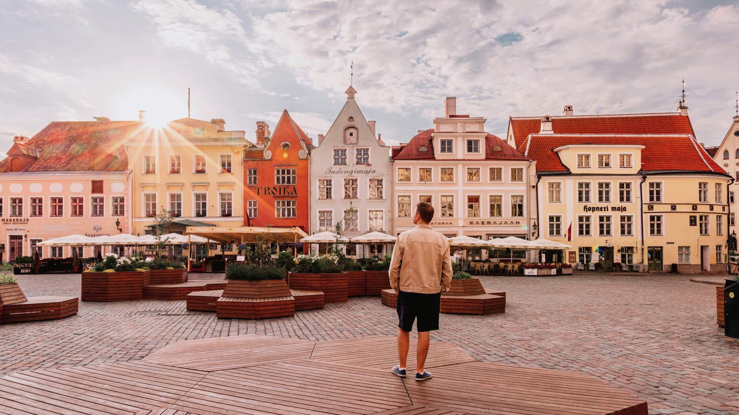 A traveller stands in a cobbled town square surrounded by colourful historic buildings at sunset in Tallinn, Estonia.