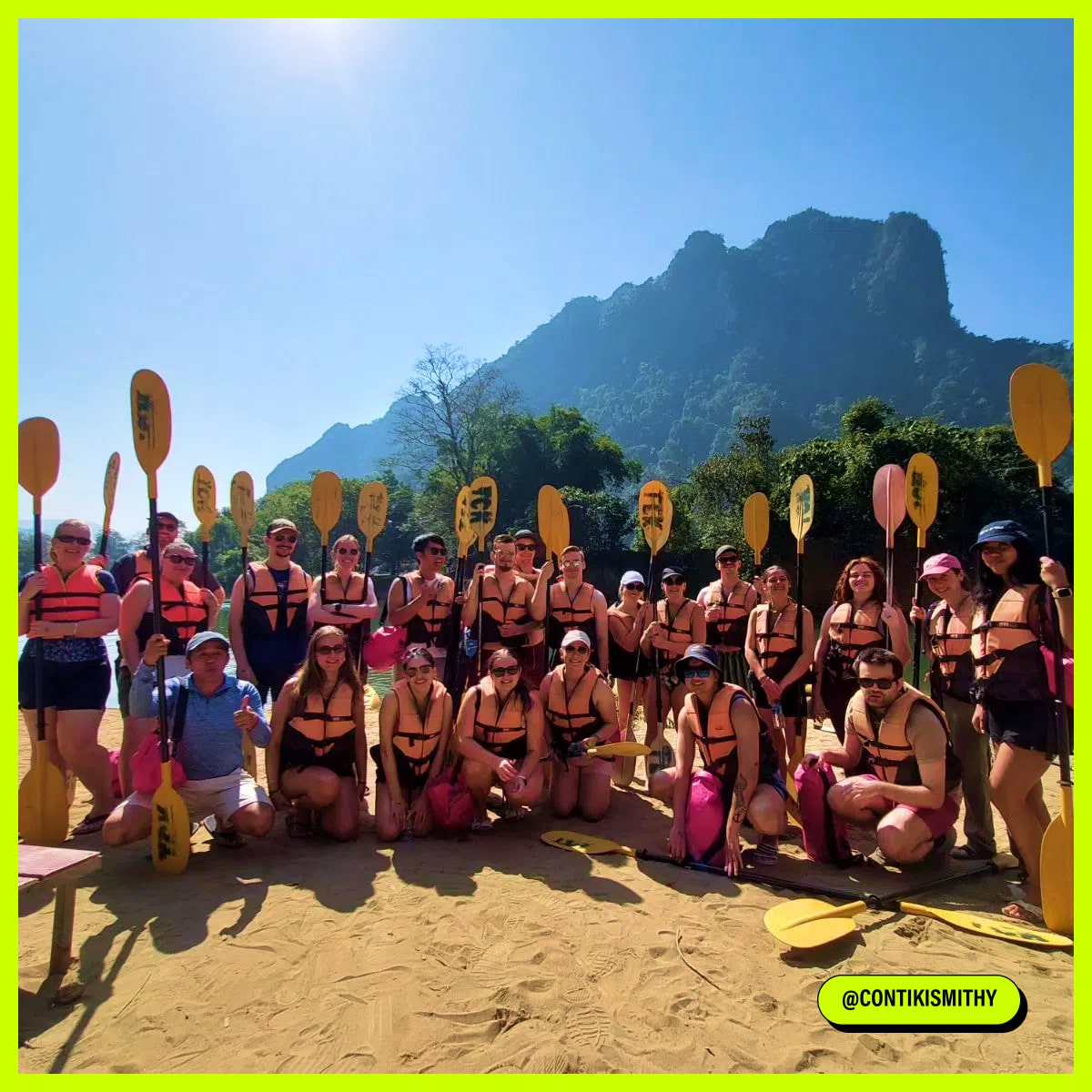 A large group of people in life jackets pose with paddles on a sunny sandy shore