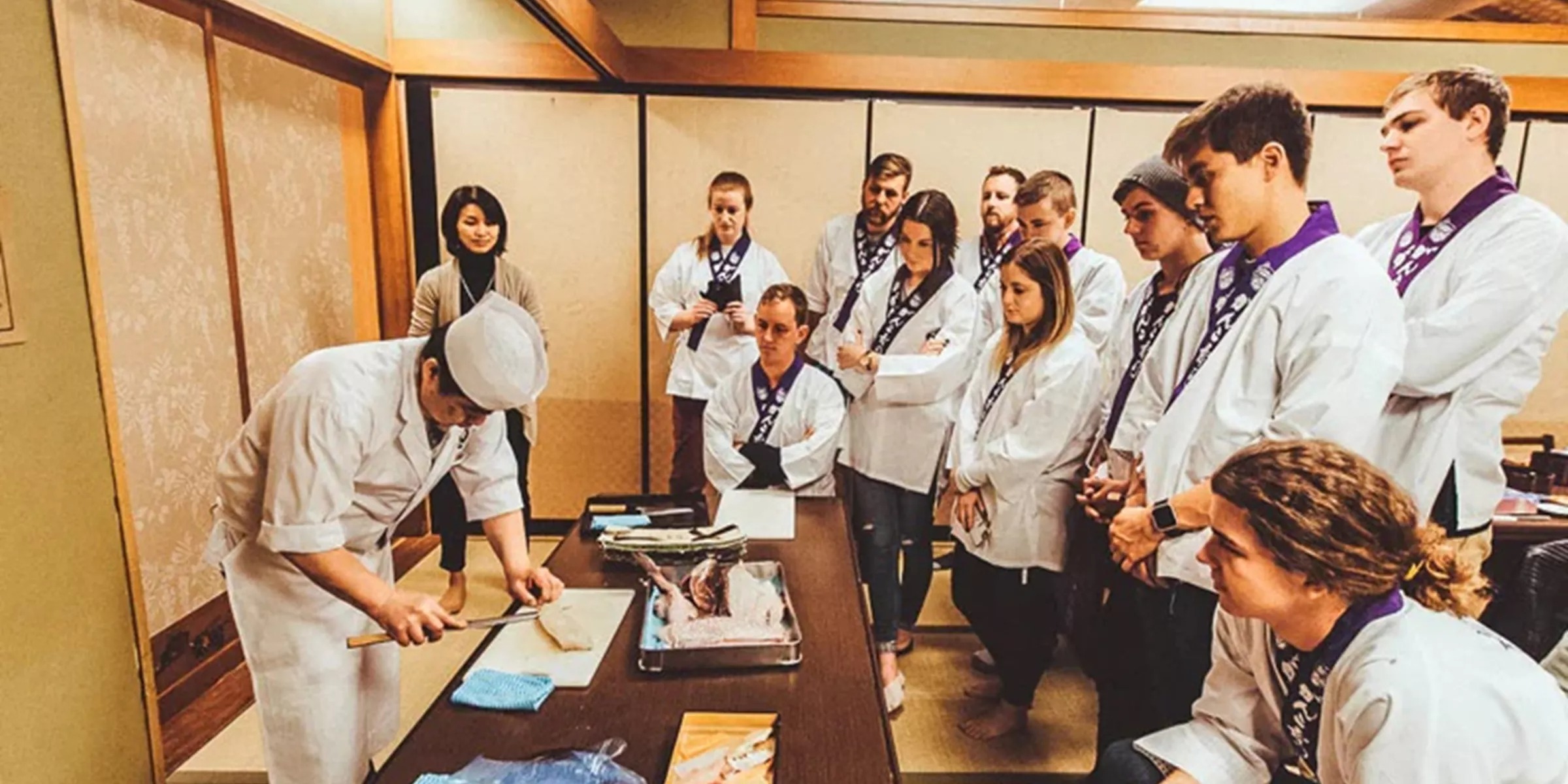 Chef cutting a slice of fish and group of students observing him during sushi cooking class