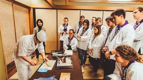 Chef cutting a slice of fish and group of students observing him during sushi cooking class