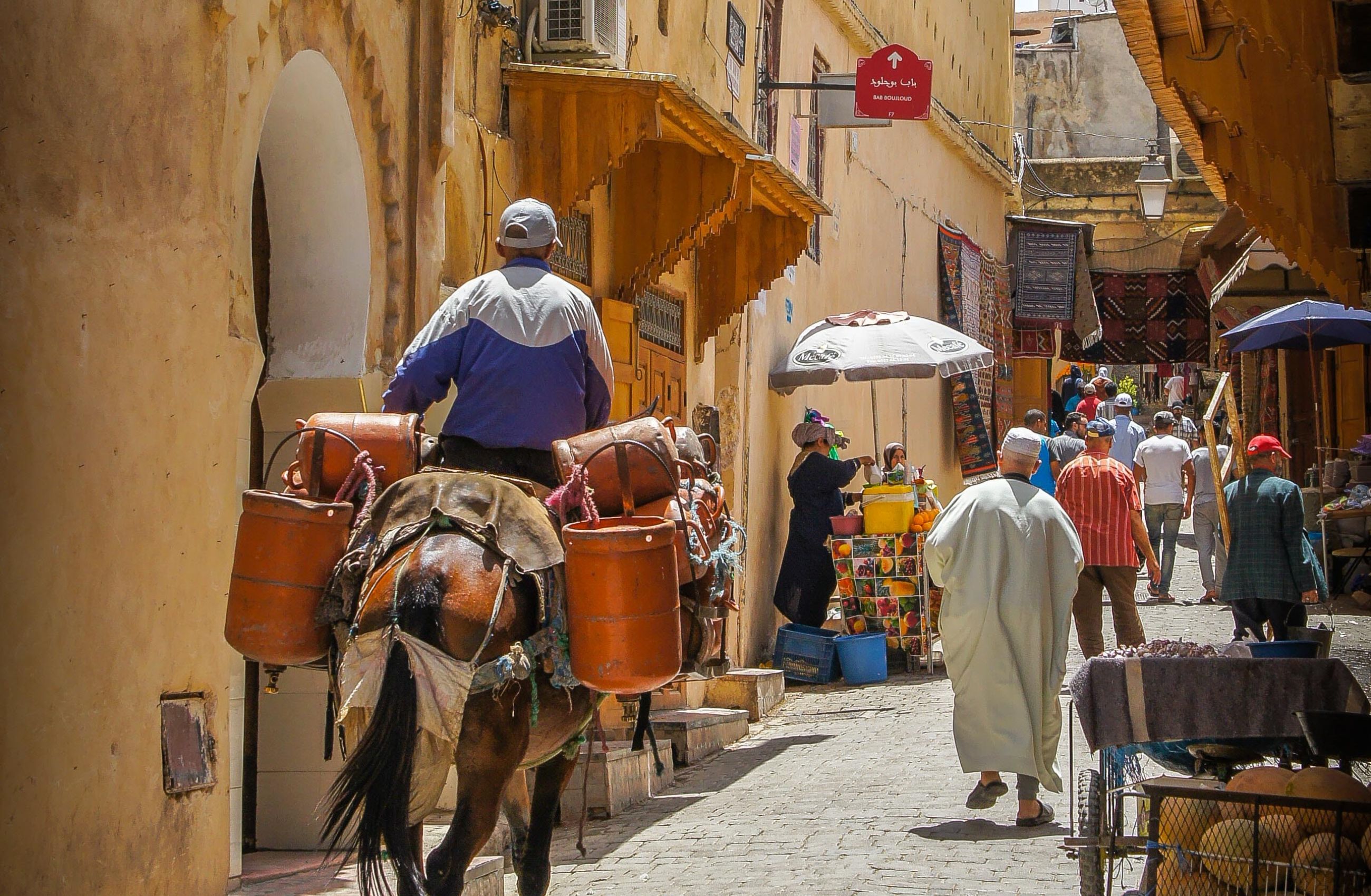 A man rides through a narrow street in Morocco on a horse. Further down the street market sellers are selling fruit and carpets.