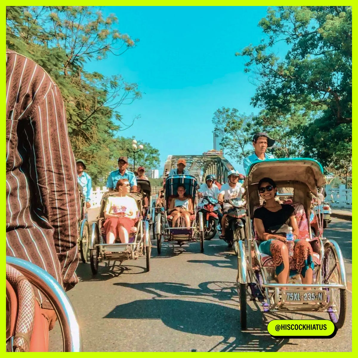 A group of people riding in cycle rickshaws on a sunny road with a bridge in the background