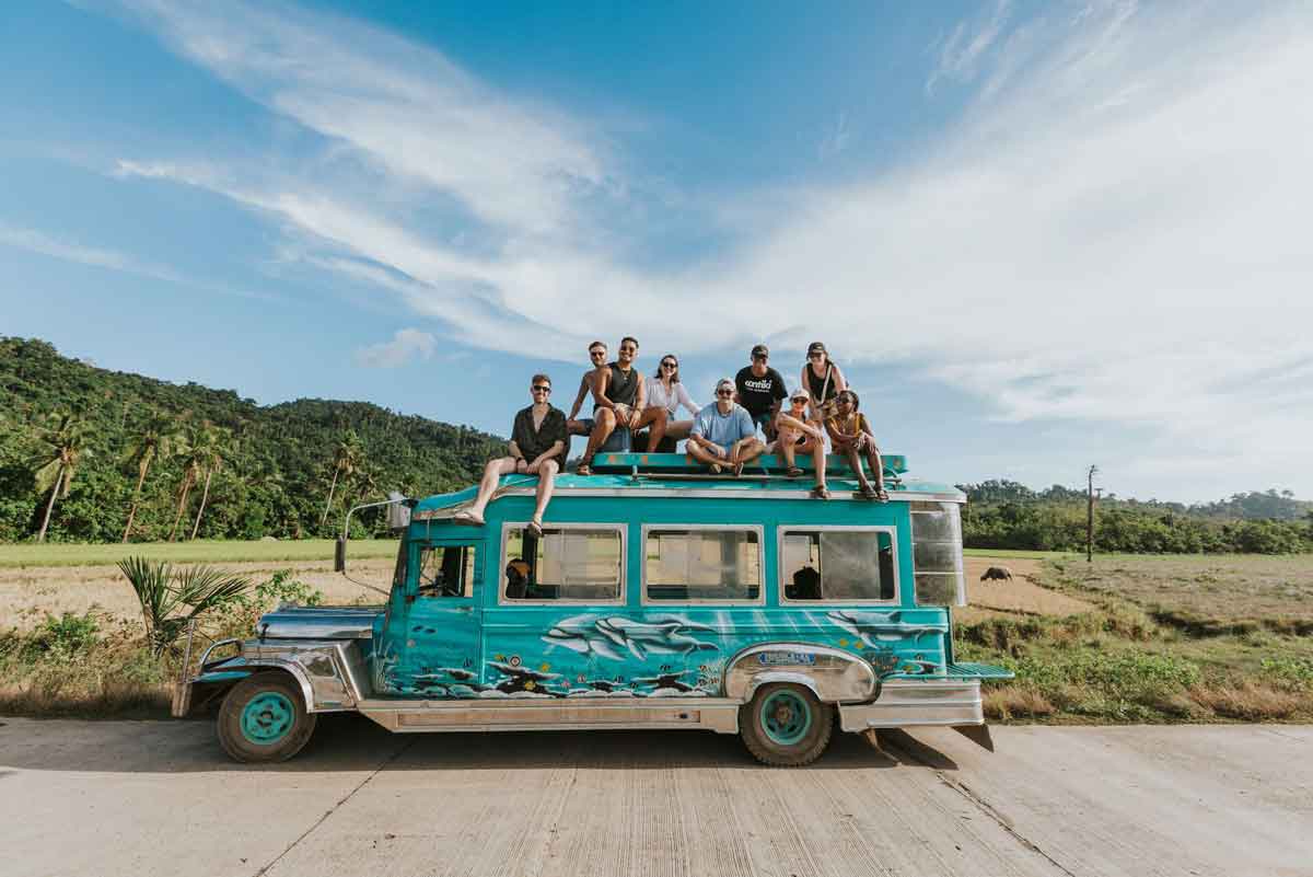 Group Of Young Friends Sitting At The Top Of A Blue Car